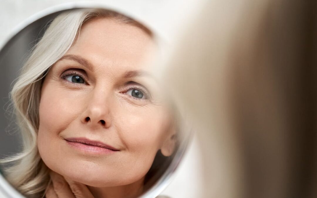 Happy 50s middle aged woman touching face skin looking in mirror reflection.